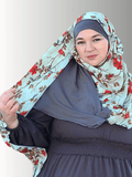 A woman showcasing a mint green hijab with striking red floral designs and a solid gray inner contrast. She is standing in front of a neutral-colored wall, gracefully holding the fabric to display its flow and intricate pattern.