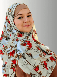 A woman wearing a mint green hijab adorned with vibrant red floral patterns,styled with a solid brown inner contrast. She is standing outdoors, surrounded by yellow autumn leaves and trees, exuding elegance and warmth in her look.