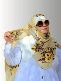 A modest golden-yellow floral hijab from the Luxe Harmony Collection, styled effortlessly with a soft white dress. The stretch chiffon fabric drapes elegantly, showcasing the vibrant blooms in a sunlit outdoor setting. Perfect for both casual and formal looks, this hijab embodies comfort and timeless elegance.