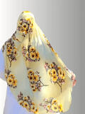 A modest golden-yellow floral hijab from the Luxe Harmony Collection, styled effortlessly with a soft white dress. The stretch chiffon fabric drapes elegantly, showcasing the vibrant blooms in a sunlit outdoor setting. Perfect for both casual and formal looks, this hijab embodies comfort and timeless elegance.