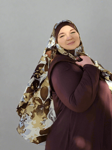 woman wearing the Golden Earth hijab outdoors on a sunny day, surrounded by autumn trees with golden leaves. The hijab showcases a soft earthy cream base with vibrant  floral patterns and delicate brown leaves, draping elegantly over her shoulder as she gazes off into the distance.